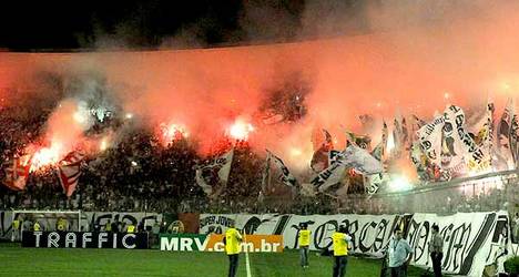 Fumo e chamas na arquibancada do Vasco, torcida com bandeiras e faixas, ambiente de estádio, clima de paixão e apoio ao time, incêndio causando preocupação entre os presentes.