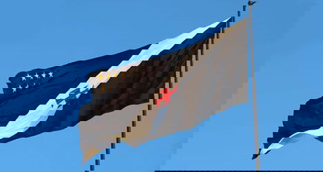 Bandeira do Vasco da Gama ao vento contra o céu azul claro, destacando as cores preta, branca, vermelha e as estrelas douradas, símbolo do clube carioca.