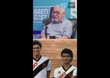 Homem falando em microfone com convidados ao fundo.