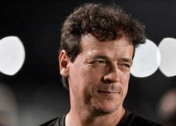 Homem sorrindo com cabelo castanho escuro e fundo de luzes brancas.