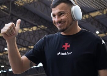 Imagem de um homem sorridente usando fones de ouvido grandes em um ambiente interno com estrutura de metal. Ele veste uma camiseta preta com logo do Betfair e o escudo do Vasco da Gama.