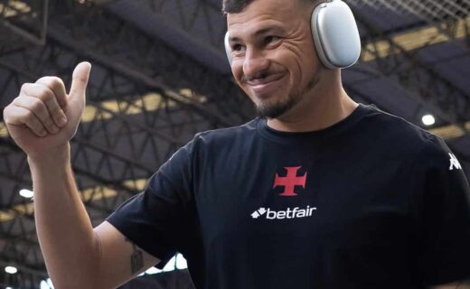 Imagem de um homem sorridente usando fones de ouvido grandes em um ambiente interno com estrutura de metal. Ele veste uma camiseta preta com logo do Betfair e o escudo do Vasco da Gama.