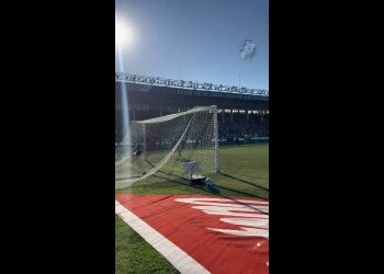 Futebol em campo com arquibancada ao fundo.