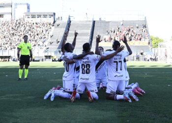 Narrador, jogadores do vasco comemorando gol em campo ao lado de torcida.