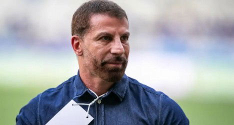 Homem de cabelo castanho, barba por fazer, usando camisa azul, em uma área de treinamento ao ar livre, com fundo desfocado de grama e céu claro.