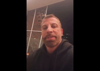 Homem com barba e hoodie escuro em ambiente interno.