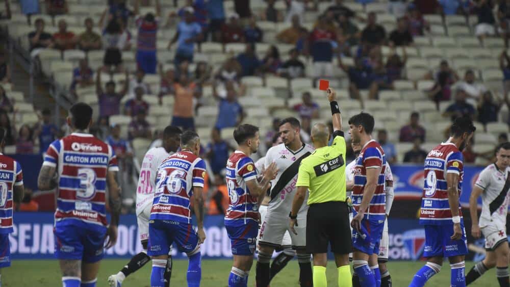 Entrada de jogadores durante o jogo no estádio do Vasco, com árbitro exibindo cartão vermelho aos jogadores. Torcida ao fundo assiste ao duelo com expectativa.