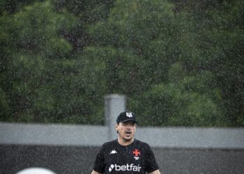 Fila de fotos do Vasco comemorando na chuva após treino, com jogador em destaque em campo ao ar livre sob chuva forte, usando uniforme preto e boné, com fundo de árvores e placas publicitárias.