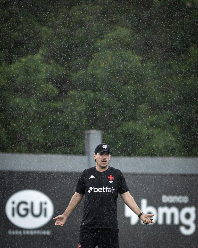 Fila de fotos do Vasco comemorando na chuva após treino, com jogador em destaque em campo ao ar livre sob chuva forte, usando uniforme preto e boné, com fundo de árvores e placas publicitárias.