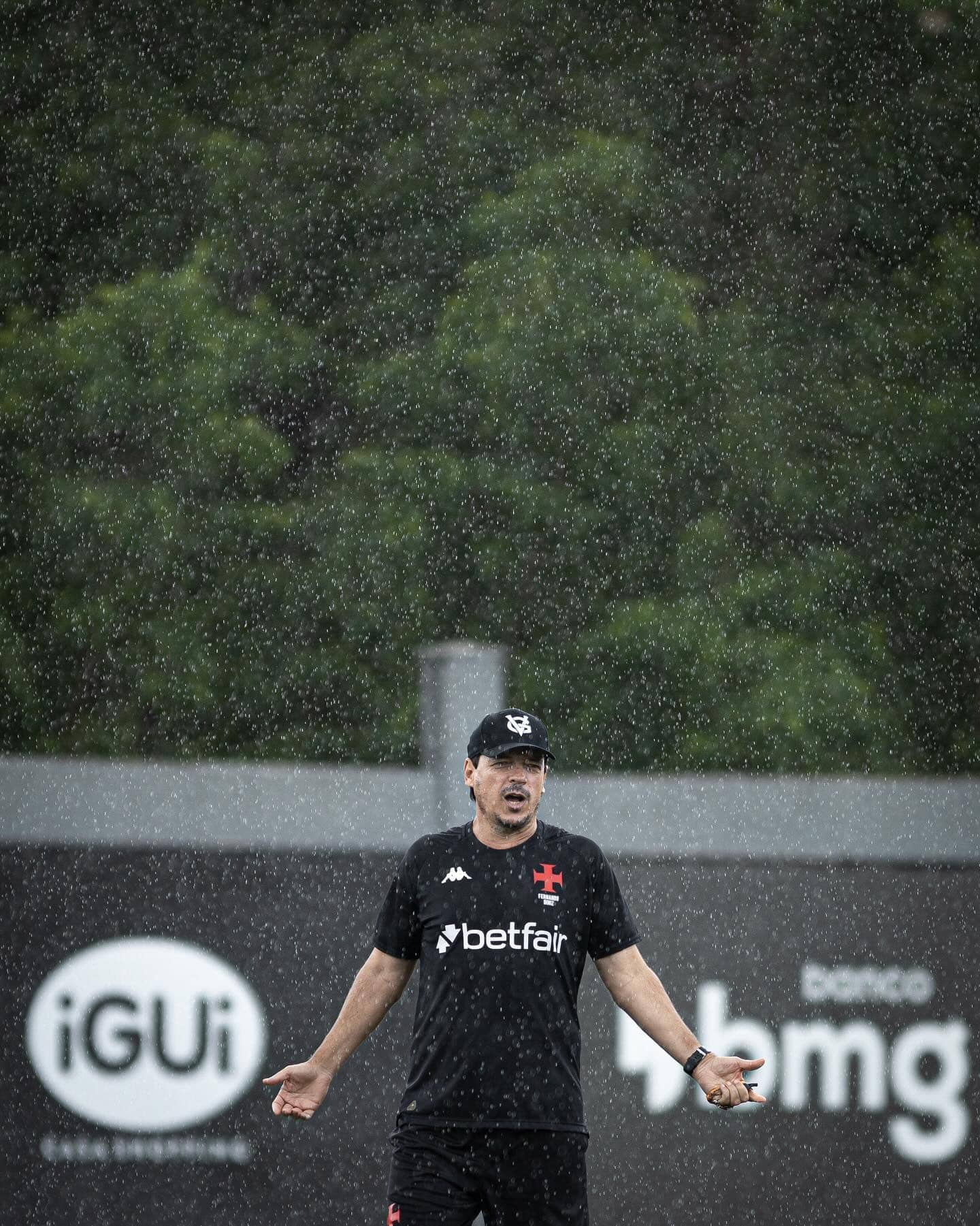 Fila de fotos do Vasco comemorando na chuva após treino, com jogador em destaque em campo ao ar livre sob chuva forte, usando uniforme preto e boné, com fundo de árvores e placas publicitárias.