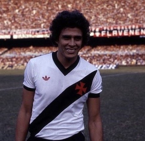 A imagem mostra um jogador do Vasco em uniforme clássico, sorrindo em um estádio cheio de torcedores vibrantes.
