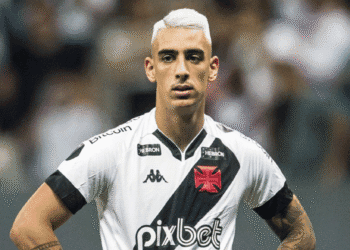 Imagem de jogador do Vasco com camisa branca e faixa preta, com expressão séria, em campo de futebol.