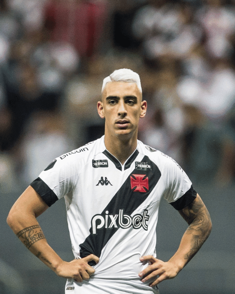 Imagem de jogador do Vasco com camisa branca e faixa preta, com expressão séria, em campo de futebol.