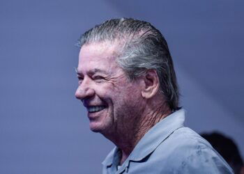 Aposentado sorrindo, cabelo grisalho, fundo azul.