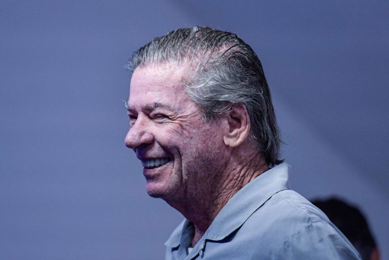 Aposentado sorrindo, cabelo grisalho, fundo azul.