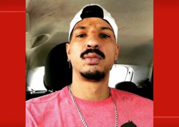 Foto de um homem com chapéu Wrangler, barba e brincos, dentro de carro, usando camiseta rosa.