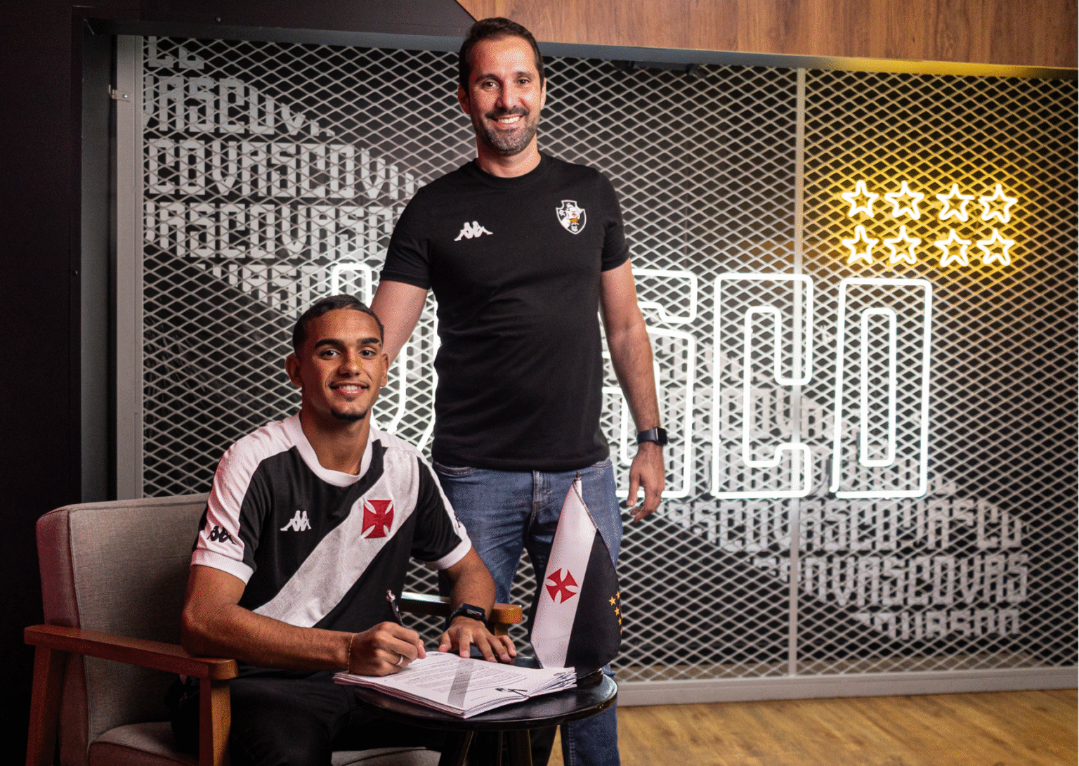 20251024-232902-2-foto-dikran-sahagian-vascocombr Afastamento de dois homens com camisetas do Vasco, um sentado assinando documento e outro de pé ao lado.