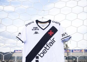 Estandarte de uma camisa do Vasco da Gama pendurada em uma rede de futebol, em um estádio de futebol ao ar livre, com grama verde e céu azul ao fundo.
