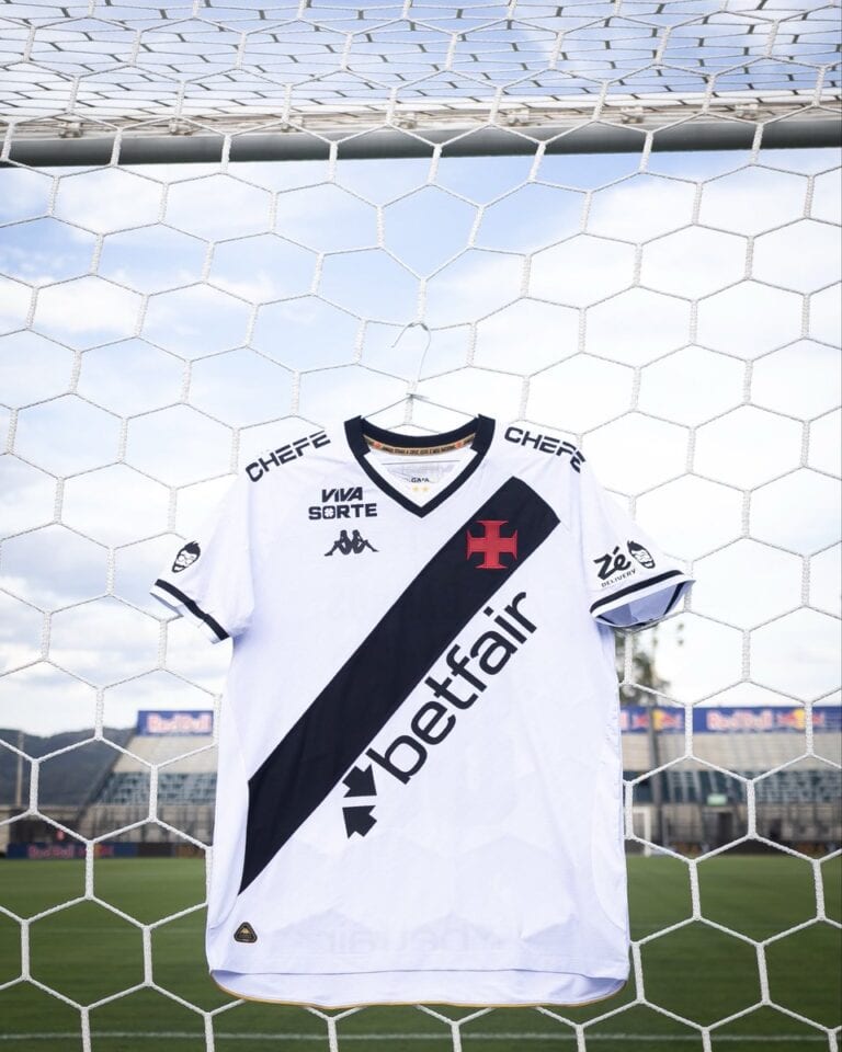 Estandarte de uma camisa do Vasco da Gama pendurada em uma rede de futebol, em um estádio de futebol ao ar livre, com grama verde e céu azul ao fundo.