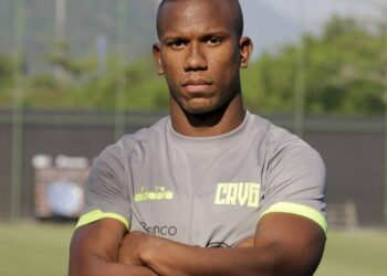 Atleta com uniforme cinza, braços cruzados, em campo de treino.