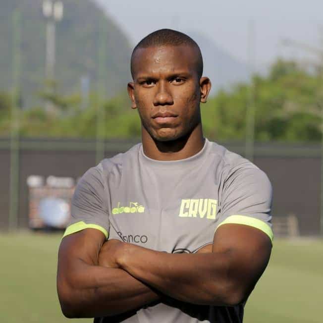 Atleta com uniforme cinza, braços cruzados, em campo de treino.