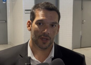 Vestido de terno preto, falando em entrevista, fundo de clínica ou consultório.