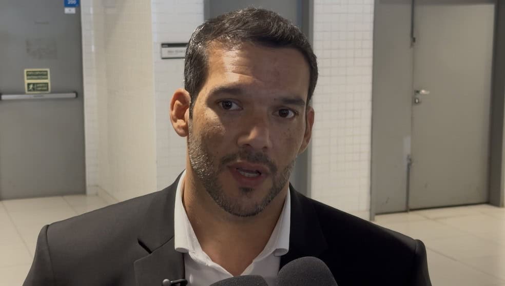 Vestido de terno preto, falando em entrevista, fundo de clínica ou consultório.