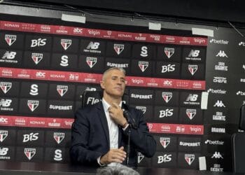 Imagem de um homem em pé, usando terno, sentado na mesa de uma coletiva de imprensa. fundo com logos do São Paulo Futebol Clube, patrocinadores e marcas associadas ao time.