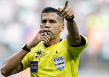 Árbitro de futebol segurando apito na boca, apontando para frente.