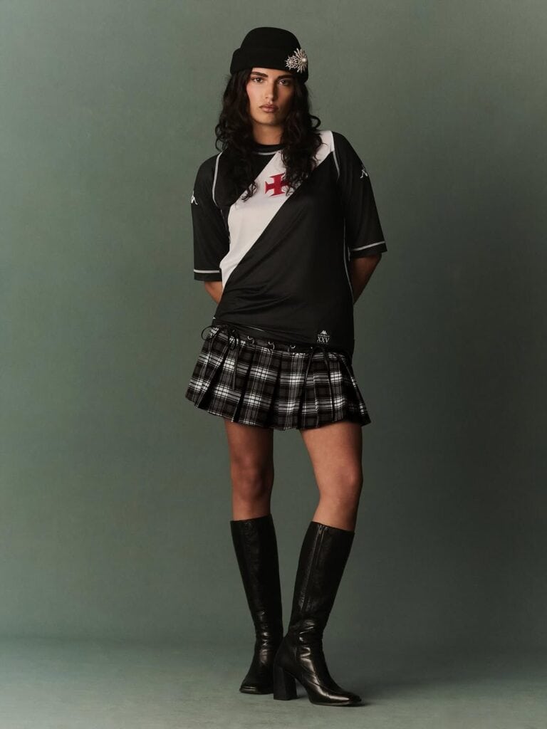 Imagem de uma mulher com uniforme de futebol, chapéu preto e bota de salto alto preta, posando com expressão séria contra fundo cinza.