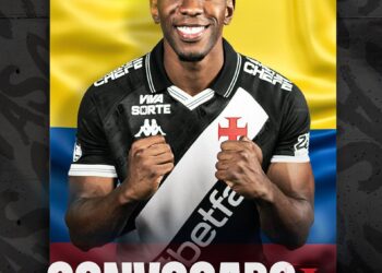 Imagem de um jogador do Vasco com uniforme, sorrindo e fazendo sinal de força com as mãos.