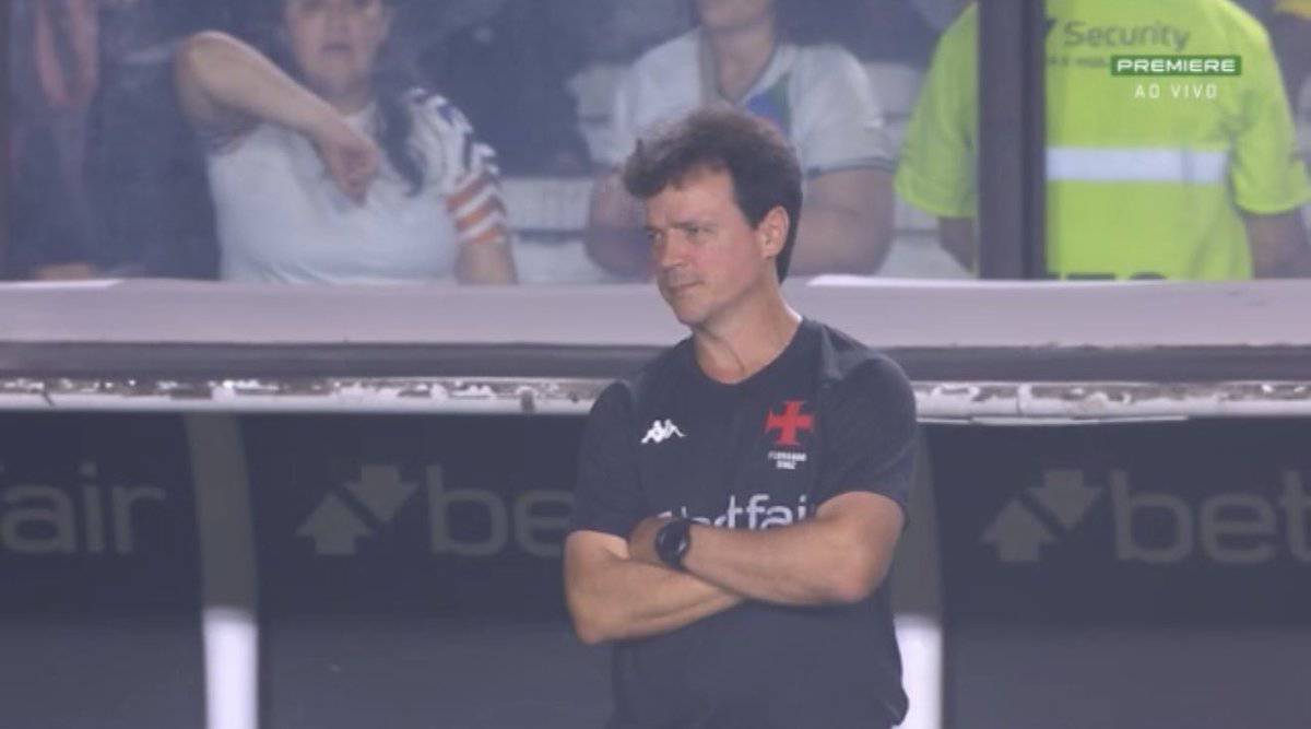 Treinador do Vasco em campo, com braços cruzados, olhar sério, usando camiseta preta com logo do clube.