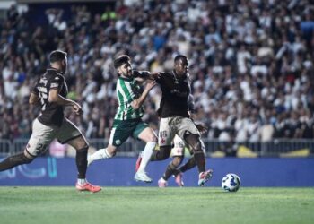 Jogadores disputam bola durante partida de futebol com torcida ao fundo.