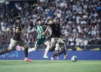 Jogadores disputam bola durante partida de futebol com torcida ao fundo.