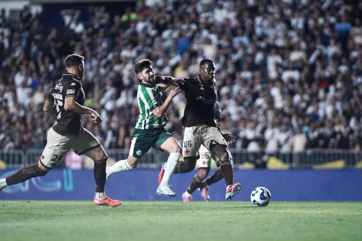 Jogadores disputam bola durante partida de futebol com torcida ao fundo.