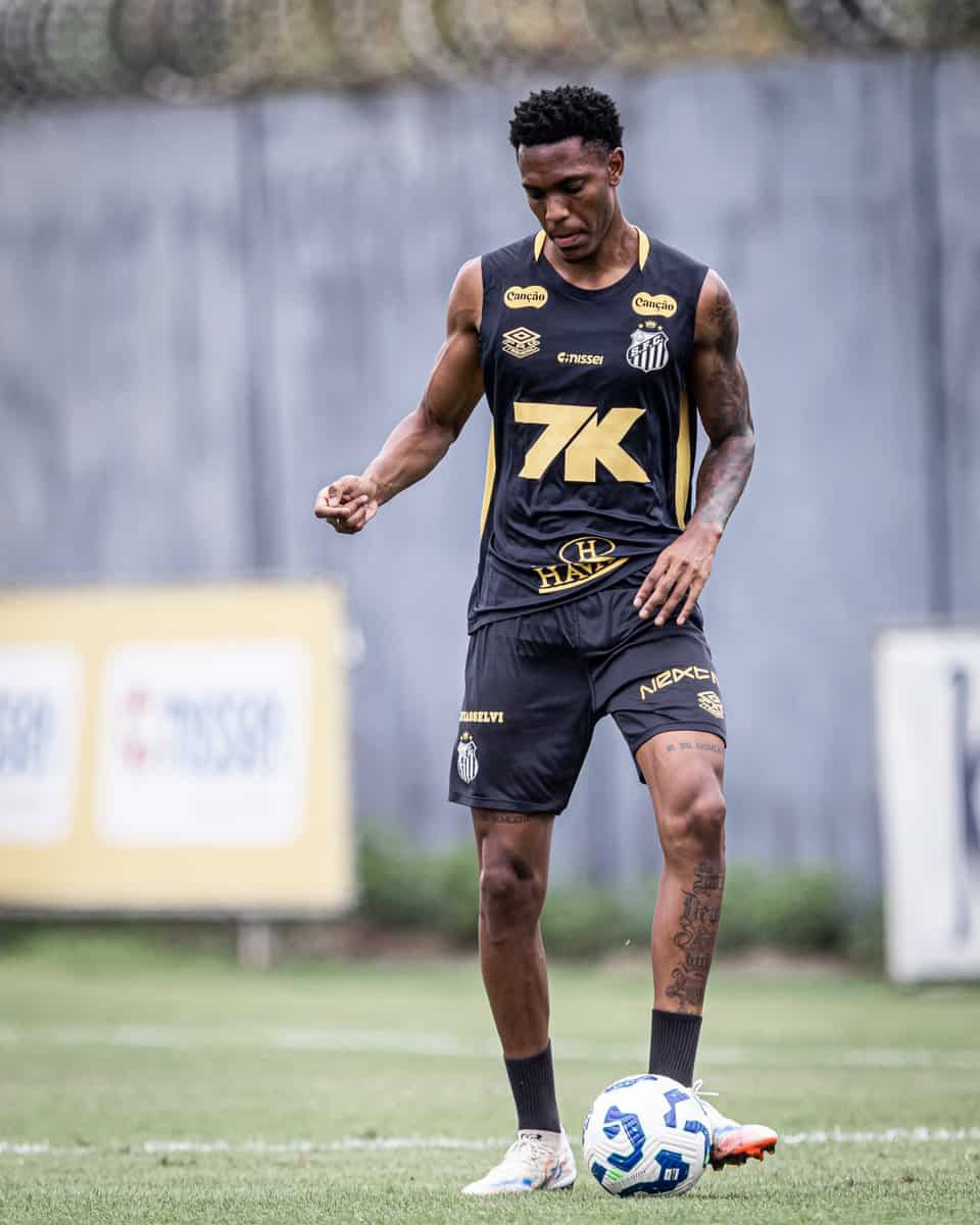 Nike jogador de futebol vestindo uniforme preto e dourado na grama com bola nos pés.