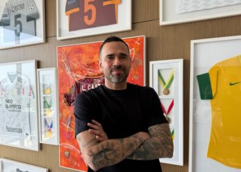 Vestido preto com braços cruzados em exibição de troféus e camisas de futebol.