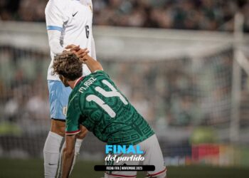 Jogador de futebol de perfil, com uniforme branco e azul, ajoelhado, apoiando a mão em um adversário no campo.