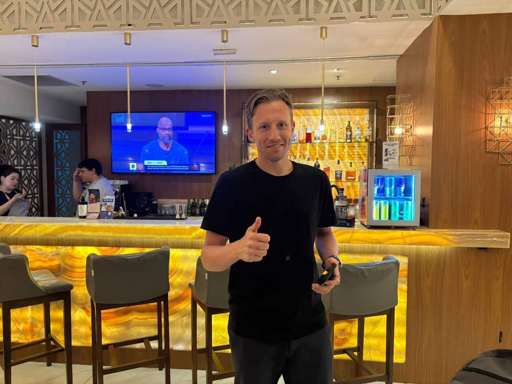 Homem sorridente em bar elegante com televisão ao fundo.