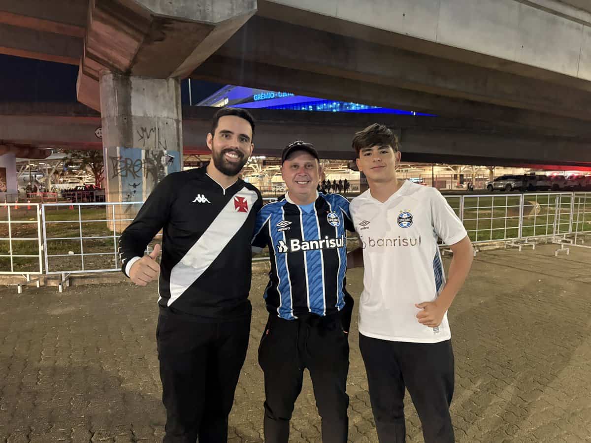 Fotógrafo com três torcedores do Grêmio, todos usando camisetas do clube, posando juntos no estádio durante a noite.