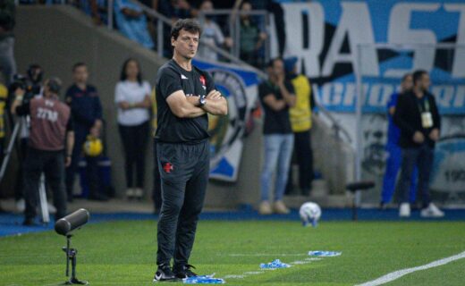 Ficha técnica do técnico do Vasco sentado no banco de reservas. Técnico atento durante partida de futebol, observando o jogo na beira do campo, com equipe e torcida ao fundo.