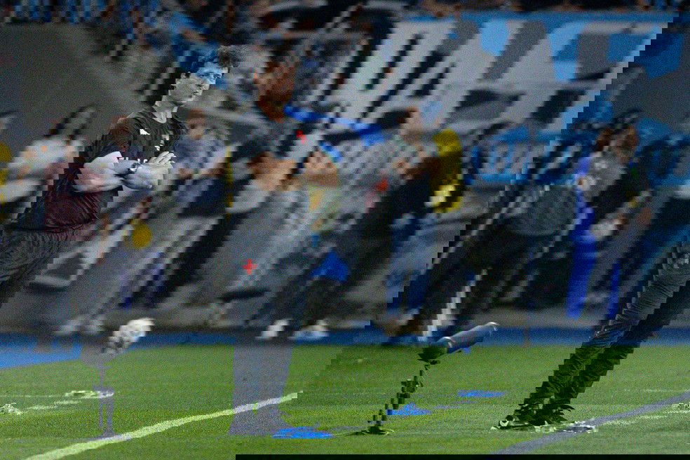 Ficha técnica do técnico do Vasco sentado no banco de reservas. Técnico atento durante partida de futebol, observando o jogo na beira do campo, com equipe e torcida ao fundo.