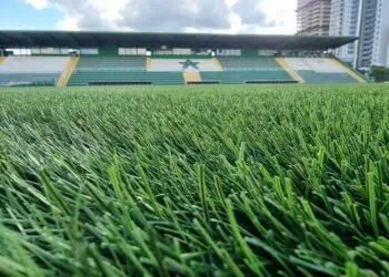 Gramado de estádio de futebol com arquibancadas ao fundo.