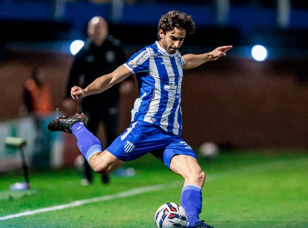 2. Jogador do Vasco em ação em campo com uniforme azul e branco, chutando uma bola com um estádio ao fundo, ao entardecer ou à noite.