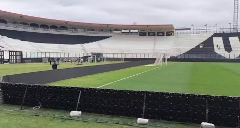 Estádio do Vasco com gramado preparado para jogo ou treino. Árbitros e equipe técnica presentes na área do campo. Arquibancadas ao fundo, quase vazias, sob céu nublado.