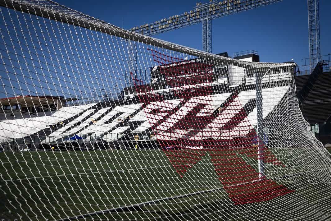 Imagem de uma rede de gol em estádio de futebol, com arquibancadas e torcedores ao fundo sob céu azul claro.