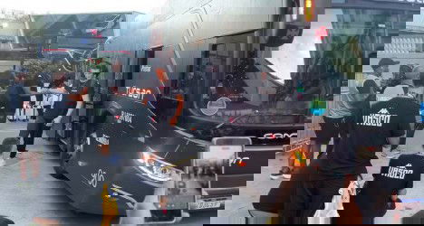 Grupo de torcedores do Vasco ao redor de um ônibus preto, alguns tirando fotos e outros conversando, na frente do veículo com detalhes visíveis na cidade.