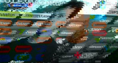 Treinador do Vasco durante entrevista após treino, com fundo de patrocinadores e ambiente esportivo ao redor.