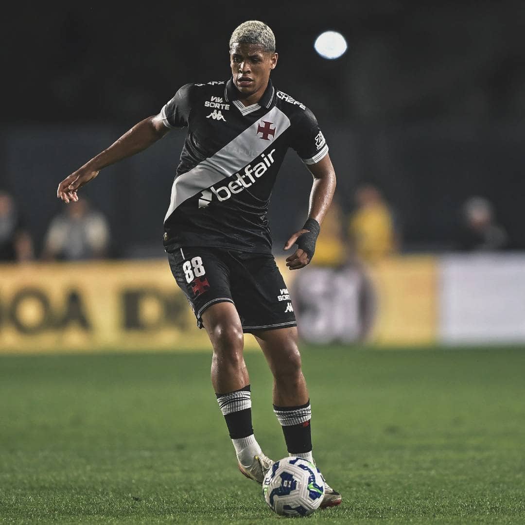 Texto alternativo: Jogador do Vasco em campo, vestido com uniforme preto e branco, driblando com a bola durante uma partida noturna, com fundo desfocado e luzes do estádio.