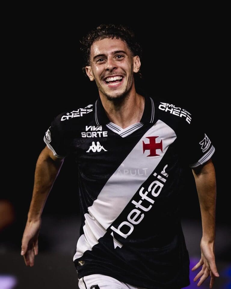 Jovem atleta com uniforme preto e branco, sorrindo com entusiasmo, celebrando vitória no estádio.